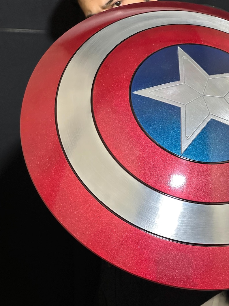 May include: A red, white, and blue shield with a silver star in the center. The shield has a circular design with concentric rings of red, silver, and blue. The shield is a replica of Captain America's shield.