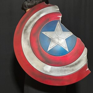 Op de afbeelding: Een Captain America-schild met een distressed design. Het schild heeft een rood-witte gestreepte rand, een zilverkleurig metalen midden en een blauwe ster in het midden. Het schild heeft een gebroken rand.