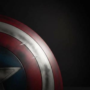 May include: Close-up of a Captain America shield. The shield features a red and silver concentric circle design with a blue star in the center. The shield has a distressed appearance with visible scratches, set against a dark background.