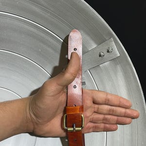May include: A silver-colored shield with concentric circular grooves. The shield has a brown leather strap with a brass buckle and a metal bracket for holding. The shield is a replica of a superhero's shield.