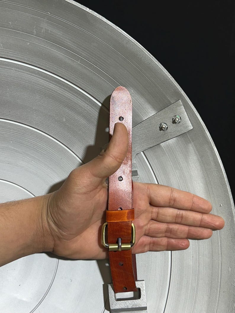 May include: A silver-colored shield with concentric circular grooves. The shield has a brown leather strap with a brass buckle and a metal bracket for holding. The shield is a replica of a superhero's shield.