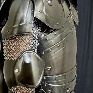 K&ouml;nnte beinhalten: Nahaufnahme eines schwarzen Metall-R&uuml;stungsteils. Die R&uuml;stung weist genietete Platten, Kettenhemden und Lederriemen auf. Die Schulter- und Ellbogensch&uuml;tzer sind gebogen und poliert. Die R&uuml;stung ist f&uuml;r den Schutz und die historische Nachstellung konzipiert.