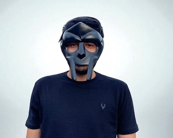 May include: Black metal mask with a distinctive design, covering the face. The mask has a dark, matte finish and features a stylized, angular shape. The person is wearing a black t-shirt.