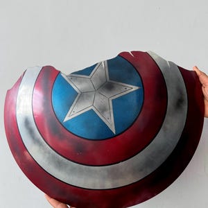May include: A damaged Captain America shield, featuring a central star design. The shield has a red, white, and blue colour scheme with a worn look. It is held against a white backdrop.