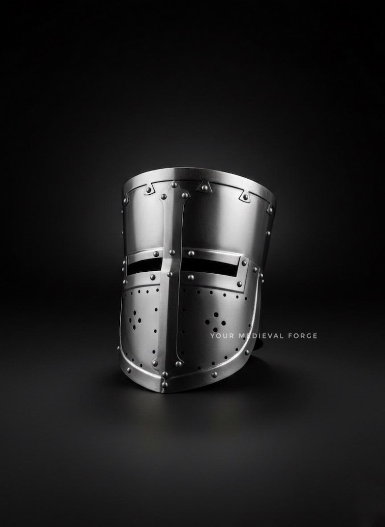 Puede incluir: Casco medieval plateado con construcci&oacute;n remachada. El casco presenta ranuras para los ojos y orificios de ventilaci&oacute;n, con el texto "YOUR MEDIEVAL FORGE" visible en la parte inferior derecha. El casco est&aacute; sobre un fondo oscuro.