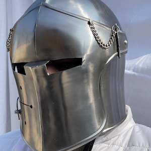 Peut inclure: Un casque m&eacute;di&eacute;val argent&eacute; et poli, avec une visi&egrave;re et des cha&icirc;nes d&eacute;coratives. Le casque pr&eacute;sente une croix sur le devant et est con&ccedil;u pour prot&eacute;ger la t&ecirc;te et le visage. Le casque est en m&eacute;tal et a une surface r&eacute;fl&eacute;chissante.