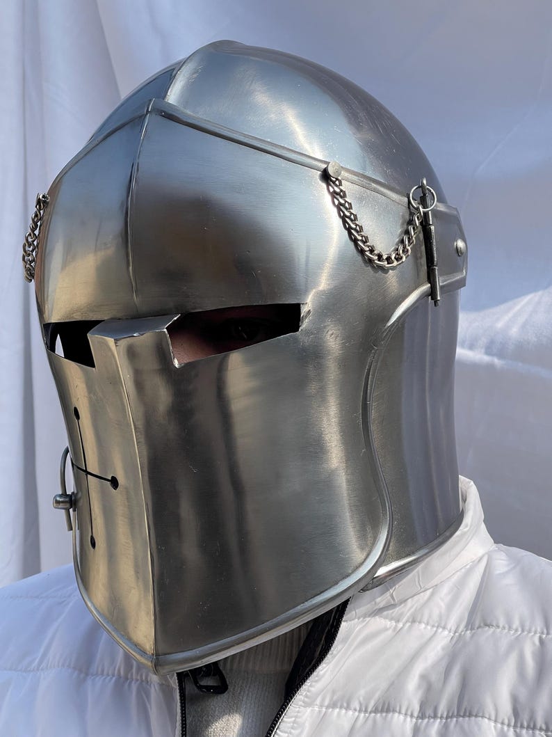 Peut inclure: Un casque m&eacute;di&eacute;val argent&eacute; et poli, avec une visi&egrave;re et des cha&icirc;nes d&eacute;coratives. Le casque pr&eacute;sente une croix sur le devant et est con&ccedil;u pour prot&eacute;ger la t&ecirc;te et le visage. Le casque est en m&eacute;tal et a une surface r&eacute;fl&eacute;chissante.