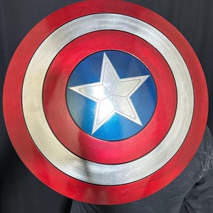 May include: A round shield with a red, white, and blue color scheme. The shield features a central blue circle with a white star, surrounded by concentric rings of silver, white, and red. The shield is a prop or collectible.