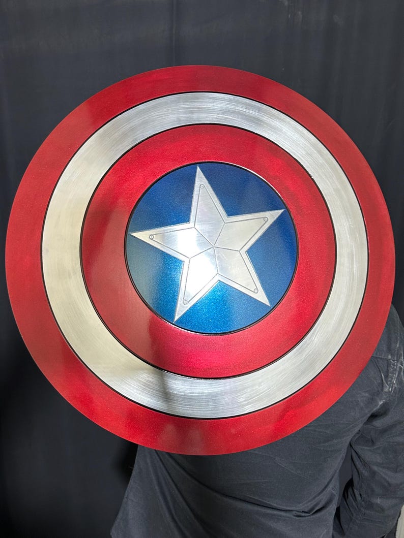 May include: A round shield with a red, white, and blue color scheme. The shield features a central blue circle with a white star, surrounded by concentric rings of silver, white, and red. The shield is a prop or collectible.