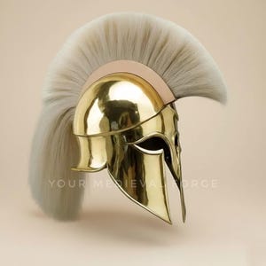 Può includere: Un elmo spartano dorato lucido con una lunga e fluente piuma grigio chiaro. L'elmo ha un design curvo con protezioni per le guance e una fascia per la fronte. Il testo "YOUR MEDIEVAL FORGE" è visibile sull'elmo.