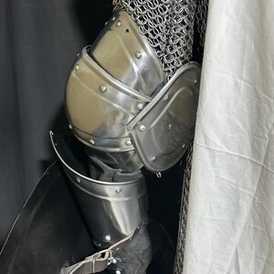 May include: Close-up of medieval armor, featuring polished steel plates and chainmail. The armor includes a breastplate, shoulder guards, and leg protection, secured with leather straps. The design suggests historical reenactment or costume use.