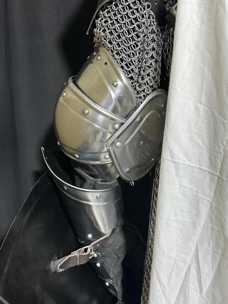 May include: Close-up of medieval armor, featuring polished steel plates and chainmail. The armor includes a breastplate, shoulder guards, and leg protection, secured with leather straps. The design suggests historical reenactment or costume use.