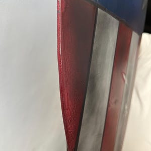 May include: A close-up of a shield with a red, white, and blue color scheme. The shield has a red border and features white stripes and a blue section with white stars. The shield appears to be made of metal.