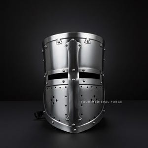 Puede incluir: Un yelmo de cruzado medieval plateado con un dise&ntilde;o de cruz. El yelmo tiene una parte superior redondeada, ranuras para los ojos y numerosos remaches. El texto "YOUR MEDIEVAL FORGE" es visible en la parte inferior derecha.