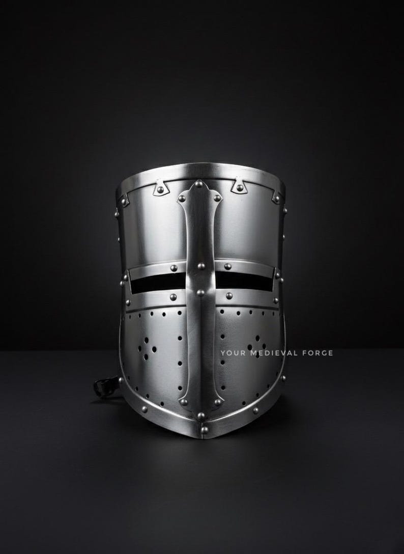 Puede incluir: Un yelmo de cruzado medieval plateado con un dise&ntilde;o de cruz. El yelmo tiene una parte superior redondeada, ranuras para los ojos y numerosos remaches. El texto "YOUR MEDIEVAL FORGE" es visible en la parte inferior derecha.