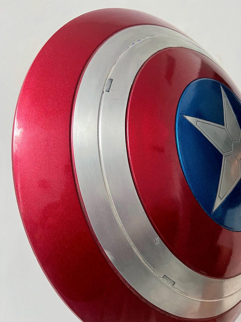 May include: A red, white, and blue Captain America shield. The shield has a metallic red finish with a silver band and a blue circle in the center. A white star is in the center of the blue circle.