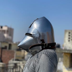 May include: A shiny steel medieval helmet with a pointed crown and visor. It is secured with a leather strap and worn over a chainmail coif. The background shows a clear blue sky and rooftops.