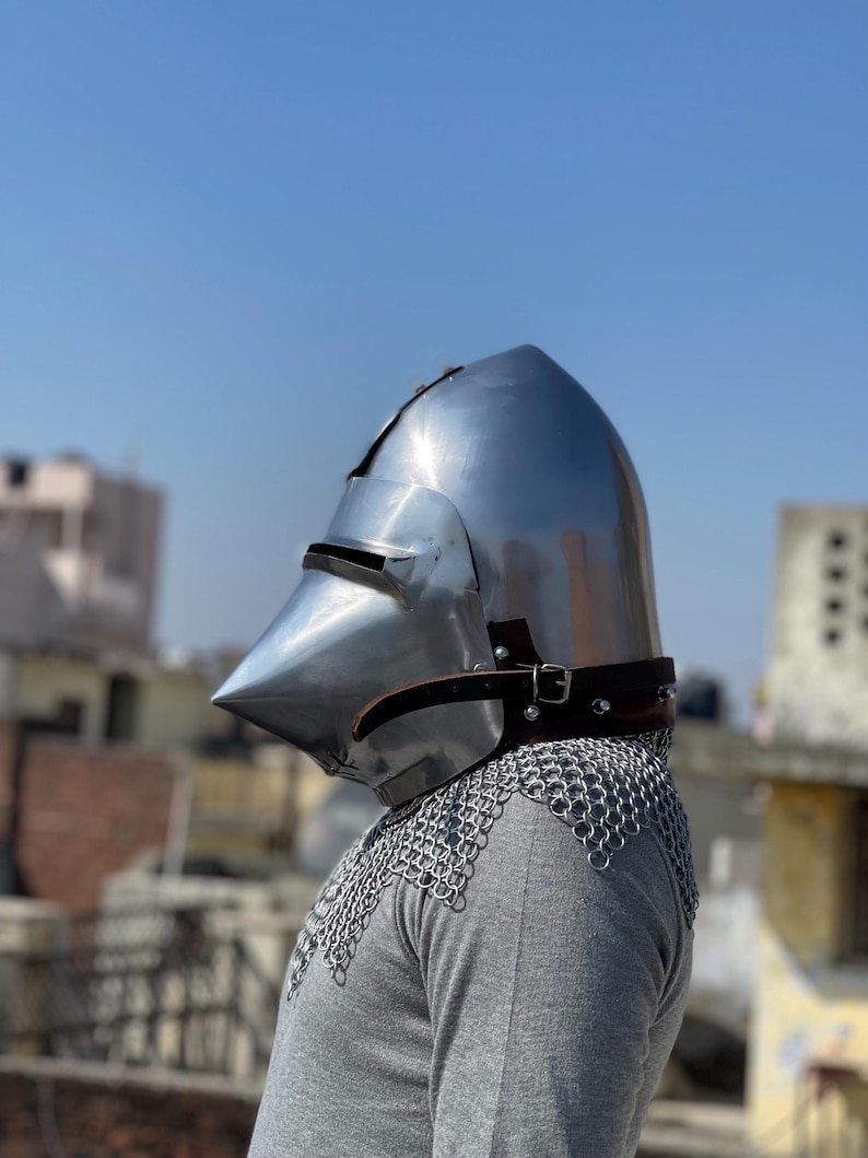 May include: A shiny steel medieval helmet with a pointed crown and visor. It is secured with a leather strap and worn over a chainmail coif. The background shows a clear blue sky and rooftops.