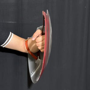 Op de afbeelding: Een zilveren en rood schild met een bruine leren riem. Het schild wordt vastgehouden door de hand van een persoon. Het schild heeft een gebogen ontwerp en een metallic afwerking.