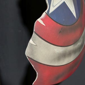 Op de afbeelding: Een beschadigd Captain America-schild, met een witte ster op een blauw veld, en rode en witte strepen. Het schild vertoont tekenen van slijtage, met krassen en beschadigingen, waardoor het een door de strijd versleten uiterlijk heeft.