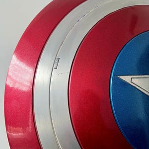 May include: A close-up of a Captain America shield. The shield features a red, white, and blue color scheme with a metallic silver band. The shield has a star emblem in the center, and the overall design is a classic superhero accessory.