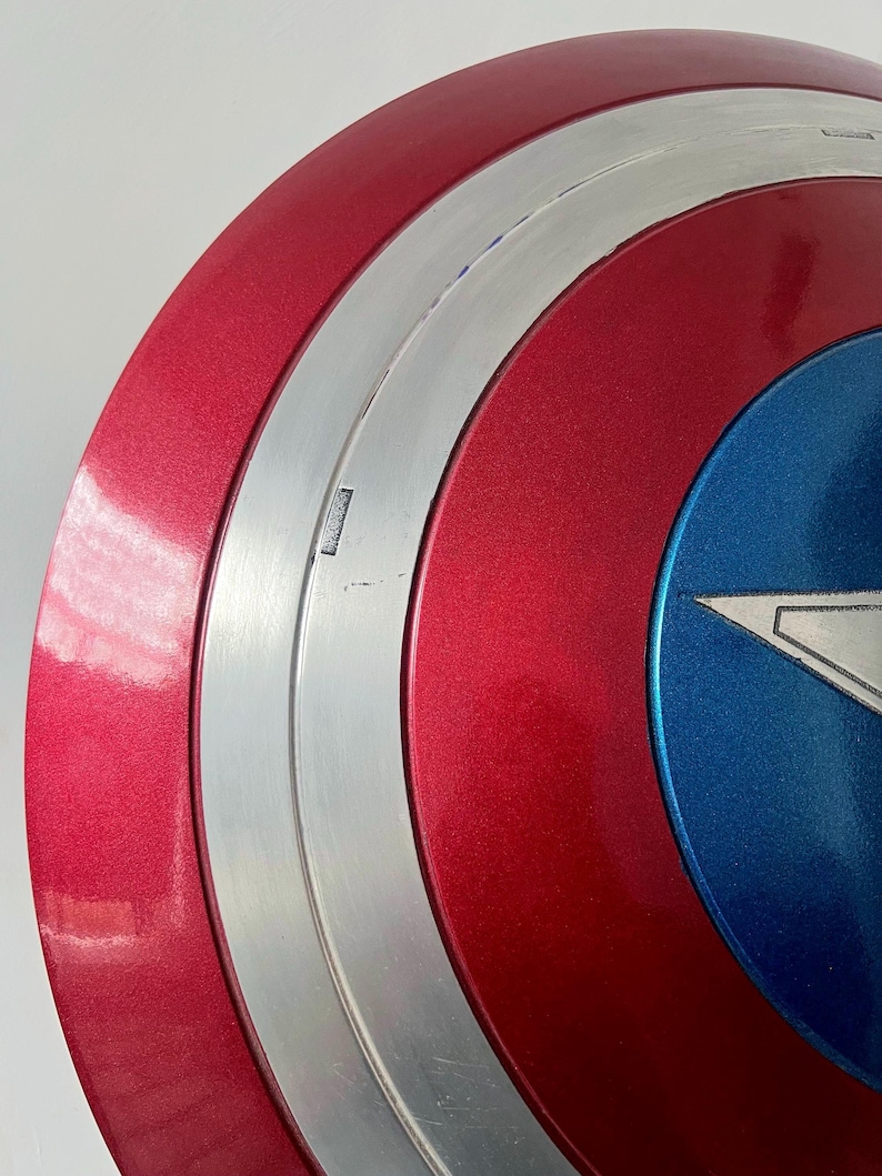 May include: A close-up of a Captain America shield. The shield features a red, white, and blue color scheme with a metallic silver band. The shield has a star emblem in the center, and the overall design is a classic superhero accessory.