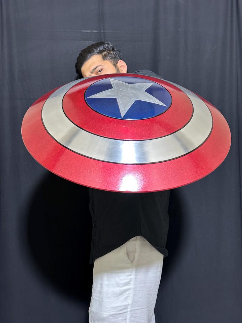 May include: A red, silver, and blue Captain America shield. The shield has a red outer ring, a silver middle ring, and a blue center with a white star. The shield is round and appears to be made of metal.