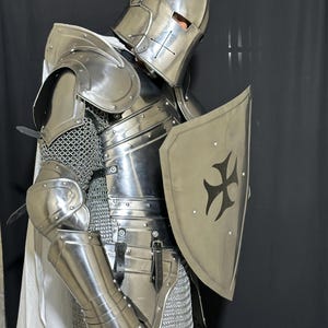 May include: A full suit of medieval knight armor, including a helmet, shoulder guards, chest plate, arm and leg protection, and a shield. The armor is silver-colored and features a cross design on the shield. A white cloth is draped over the shoulders.