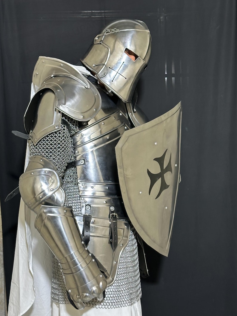 May include: A full suit of medieval knight armor, including a helmet, shoulder guards, chest plate, arm and leg protection, and a shield. The armor is silver-colored and features a cross design on the shield. A white cloth is draped over the shoulders.