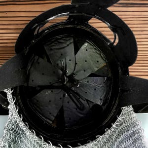 Puede incluir: Casco de metal negro con protector de cuello de cota de malla. El interior presenta un forro de cuero con un dise&ntilde;o de estrella. El casco tiene una visera curva y est&aacute; sobre un fondo de madera.
