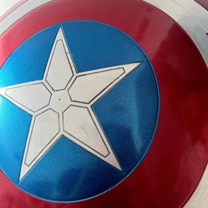 May include: A close-up of a Captain America shield. The shield features a red and silver outer ring, a blue center with a white star, and a glossy finish. The shield is a replica of the iconic superhero prop.