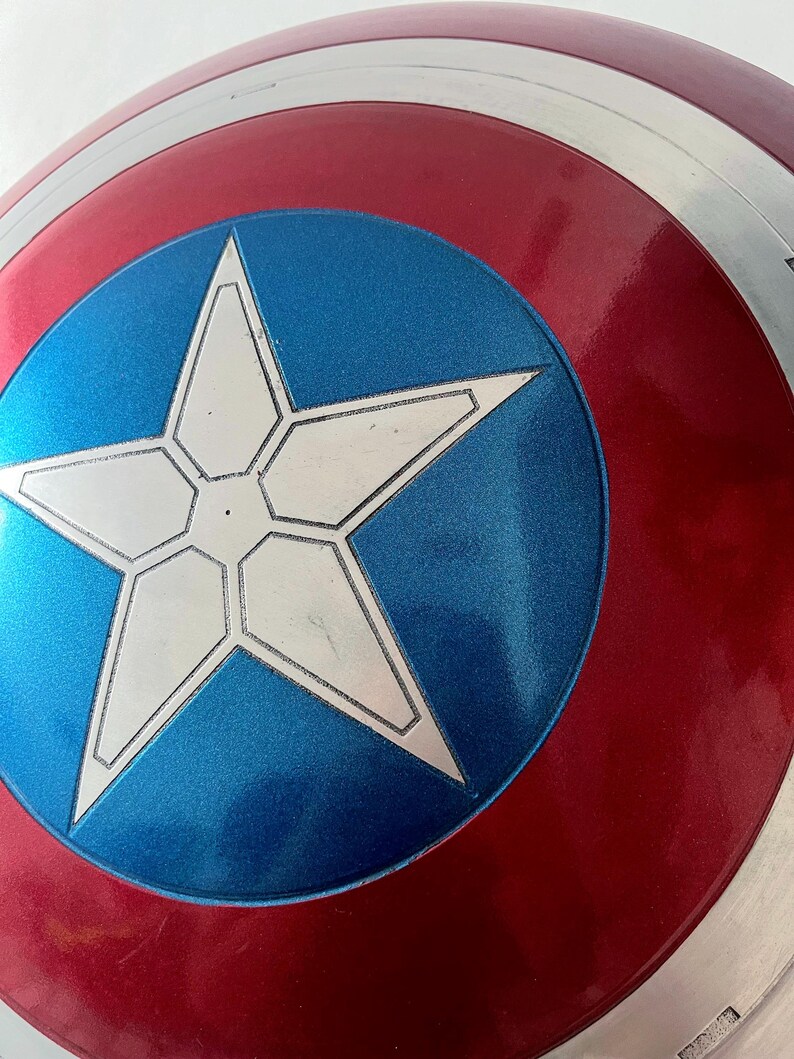 May include: A close-up of a Captain America shield. The shield features a red and silver outer ring, a blue center with a white star, and a glossy finish. The shield is a replica of the iconic superhero prop.