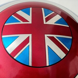May include: A metallic red shield with a Union Jack design. The shield features a red outer ring, a silver inner ring, and a central design with red, white, and blue elements. The shield is a replica of a superhero's shield.
