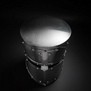 Puede incluir: Un casco medieval plateado con la parte superior redondeada y construcci&oacute;n remachada. El casco presenta una visera con orificios de ventilaci&oacute;n, sobre un fondo oscuro. El texto "YOUR MEDIEVAL FORGE" es visible.