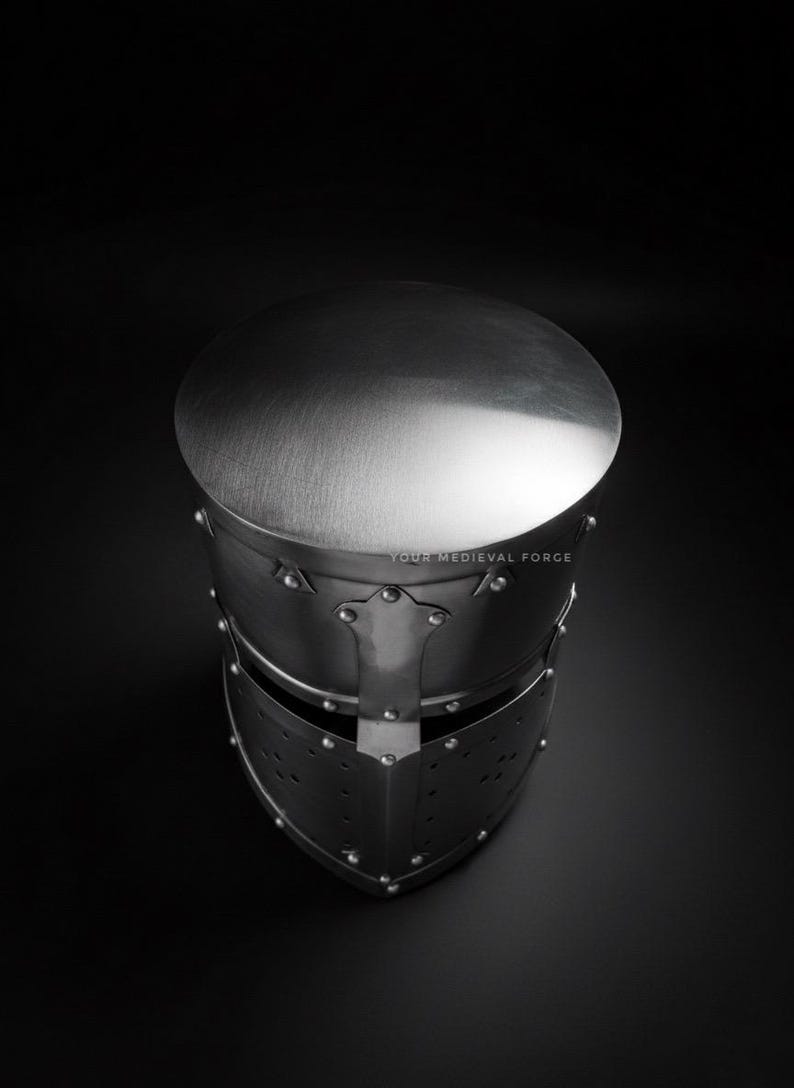 Puede incluir: Un casco medieval plateado con la parte superior redondeada y construcci&oacute;n remachada. El casco presenta una visera con orificios de ventilaci&oacute;n, sobre un fondo oscuro. El texto "YOUR MEDIEVAL FORGE" es visible.