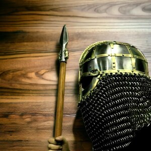 Puede incluir: Un casco de estilo medieval con una protecci&oacute;n facial de cota de malla y una lanza. El casco es de metal con detalles dorados. La lanza tiene un asta de madera y una punta de lanza de metal. Fondo de madera.
