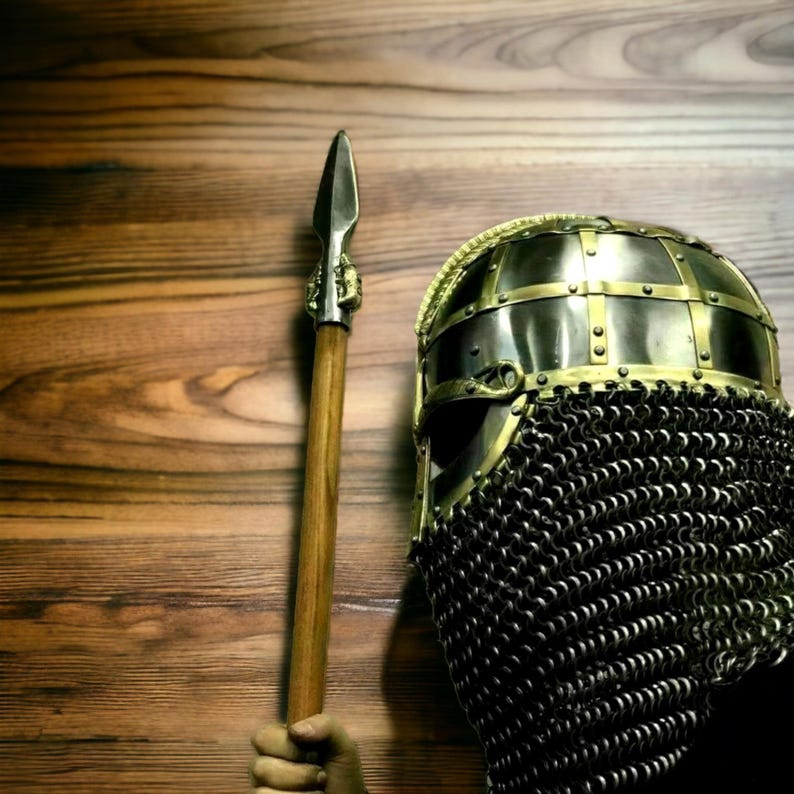 Puede incluir: Un casco de estilo medieval con una protecci&oacute;n facial de cota de malla y una lanza. El casco es de metal con detalles dorados. La lanza tiene un asta de madera y una punta de lanza de metal. Fondo de madera.