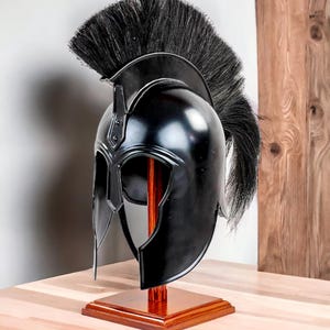 Könnte beinhalten: Schwarzer spartanischer Helm mit schwarzer Feder, präsentiert auf einem Holzständer. Der Helm hat eine polierte Oberfläche und detaillierte Gesichtsmerkmale. Der Ständer besteht aus Holz und hat eine quadratische Basis.