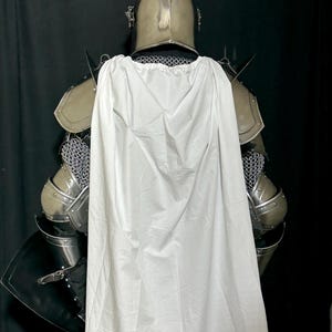May include: A full suit of medieval armor, including a helmet, shoulder guards, and chainmail. A white cape is draped over the back of the armor. The armor is silver-colored, and the cape is white.