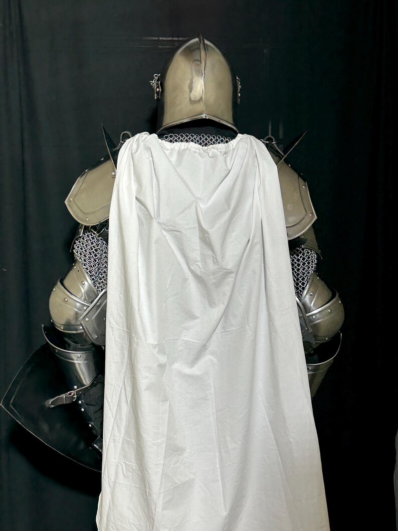 May include: A full suit of medieval armor, including a helmet, shoulder guards, and chainmail. A white cape is draped over the back of the armor. The armor is silver-colored, and the cape is white.
