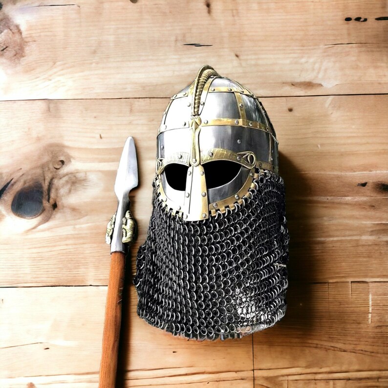 Puede incluir: Un casco de estilo vikingo con una protecci&oacute;n facial de cota de malla y detalles en lat&oacute;n. Una lanza con un asta de madera y una punta de lanza de metal se encuentra junto al casco. Sobre una superficie de madera.