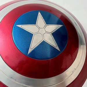 May include: A red, white, and blue shield with a star emblem. The shield has a metallic silver border and a glossy finish. The central emblem is a white star on a blue circle.