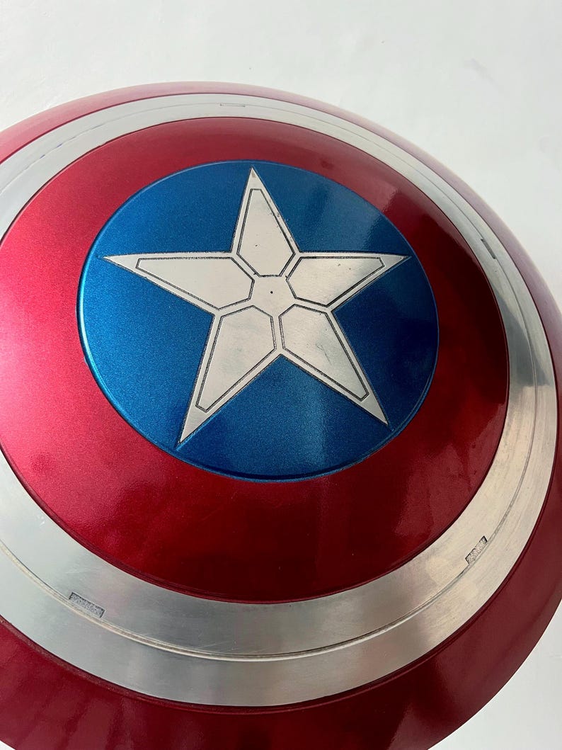 May include: A red, white, and blue shield with a star emblem. The shield has a metallic silver border and a glossy finish. The central emblem is a white star on a blue circle.