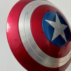 May include: A red, white, and blue Captain America shield. The shield has a metallic finish with a silver ring and a blue center featuring a white star. The shield is a replica of the iconic superhero prop.