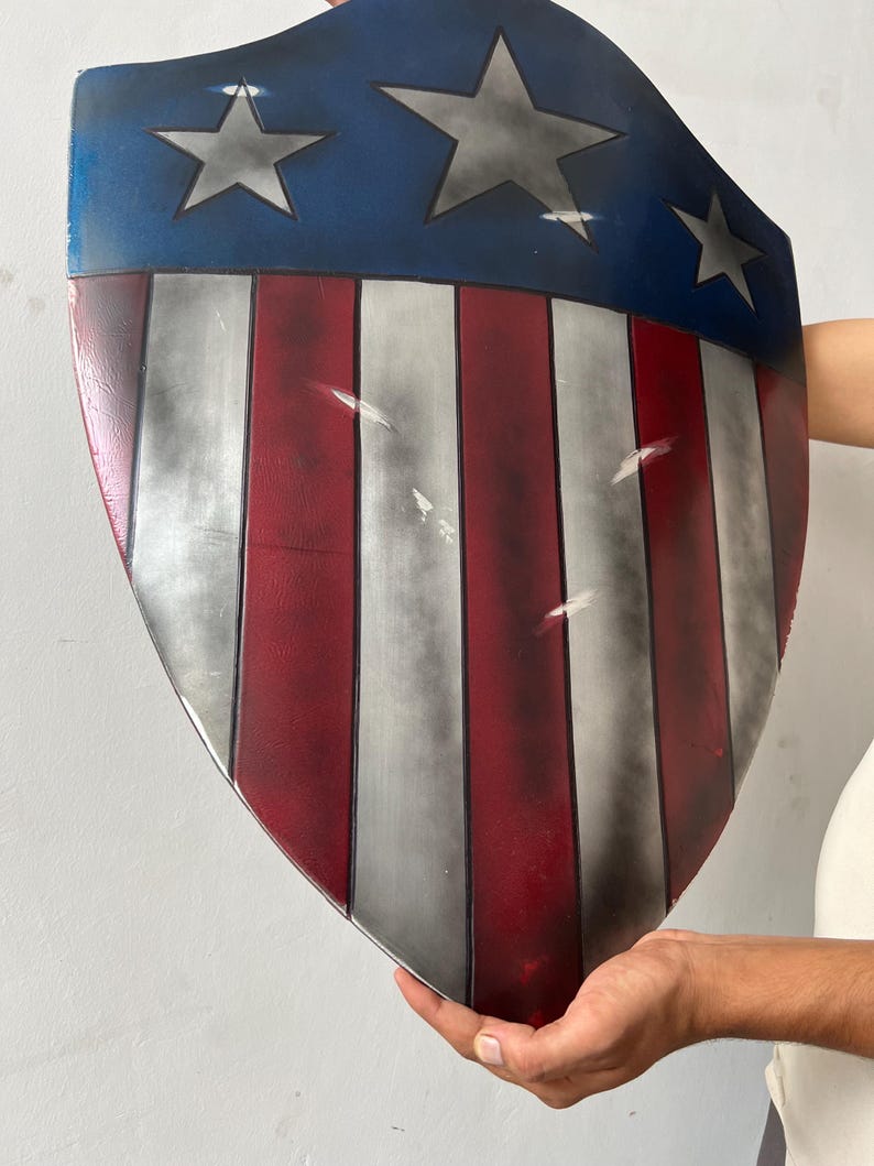 May include: A shield with a blue top section featuring three gray stars. The shield has red and white vertical stripes. The shield is held by a person.