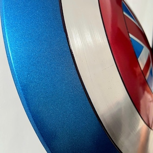 May include: A metallic blue, red, white, and silver shield with a star and stripes design. The shield has a circular shape with a blue outer rim and a central design featuring red, white, and blue elements.
