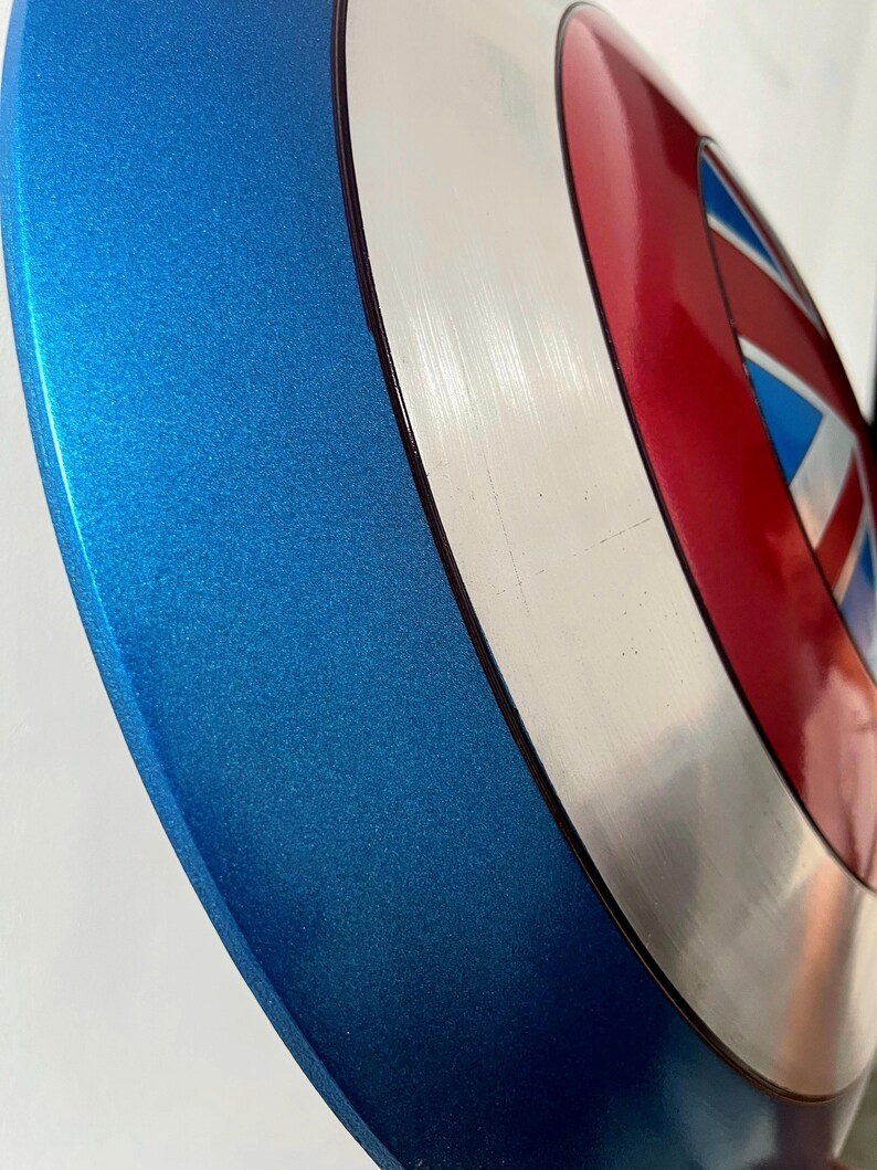 May include: A metallic blue, red, white, and silver shield with a star and stripes design. The shield has a circular shape with a blue outer rim and a central design featuring red, white, and blue elements.