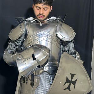 May include: A full suit of polished steel armor, including a helmet, chest plate, shoulder guards, and chainmail. A triangular shield with a black cross design is also present. The armor is designed to protect the wearer.