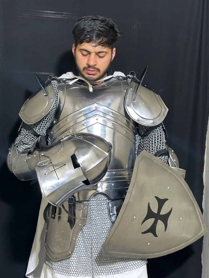 May include: A full suit of polished steel armor, including a helmet, chest plate, shoulder guards, and chainmail. A triangular shield with a black cross design is also present. The armor is designed to protect the wearer.