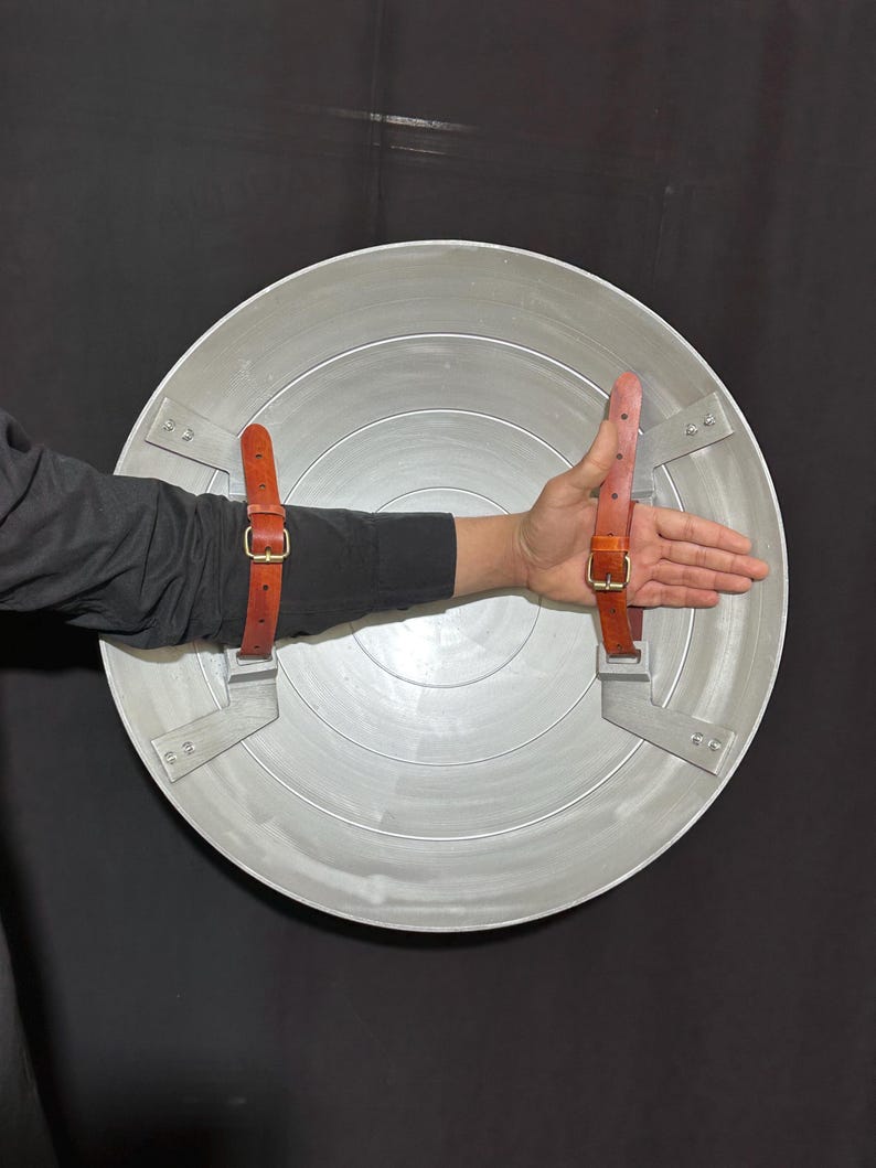 May include: A silver-colored, circular shield with concentric rings and brown leather straps. The shield has a metallic appearance and is designed with a series of rivets and mounting brackets. The straps are secured with gold-colored buckles.